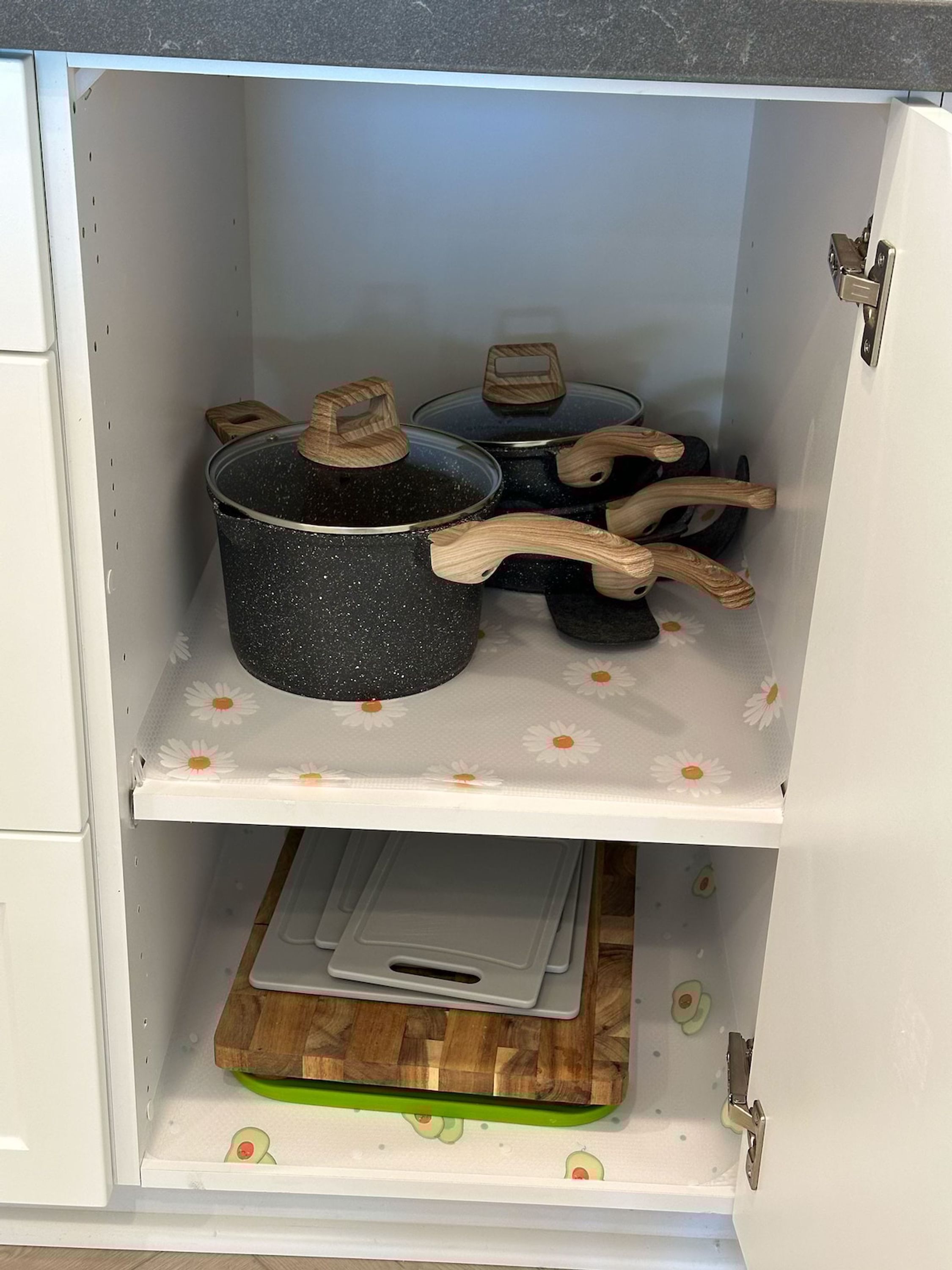 Un mueble de cocina con tres ollas negras en el estante superior y una tabla de cortar en el inferior.