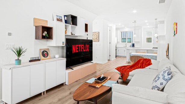 Een moderne woonkamer met een flatscreen-tv, witte meubels en een keuken op de achtergrond.