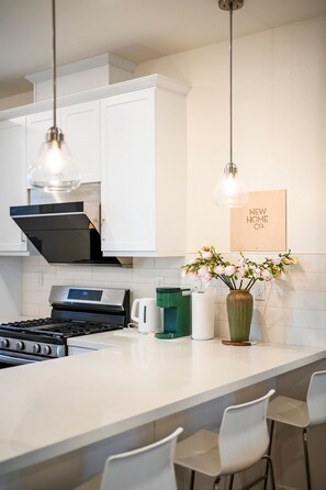 A modern kitchen with white cabinetry, stainless steel appliances, a central island with bar stools, pendant lighting, and a framed sign on the wall.