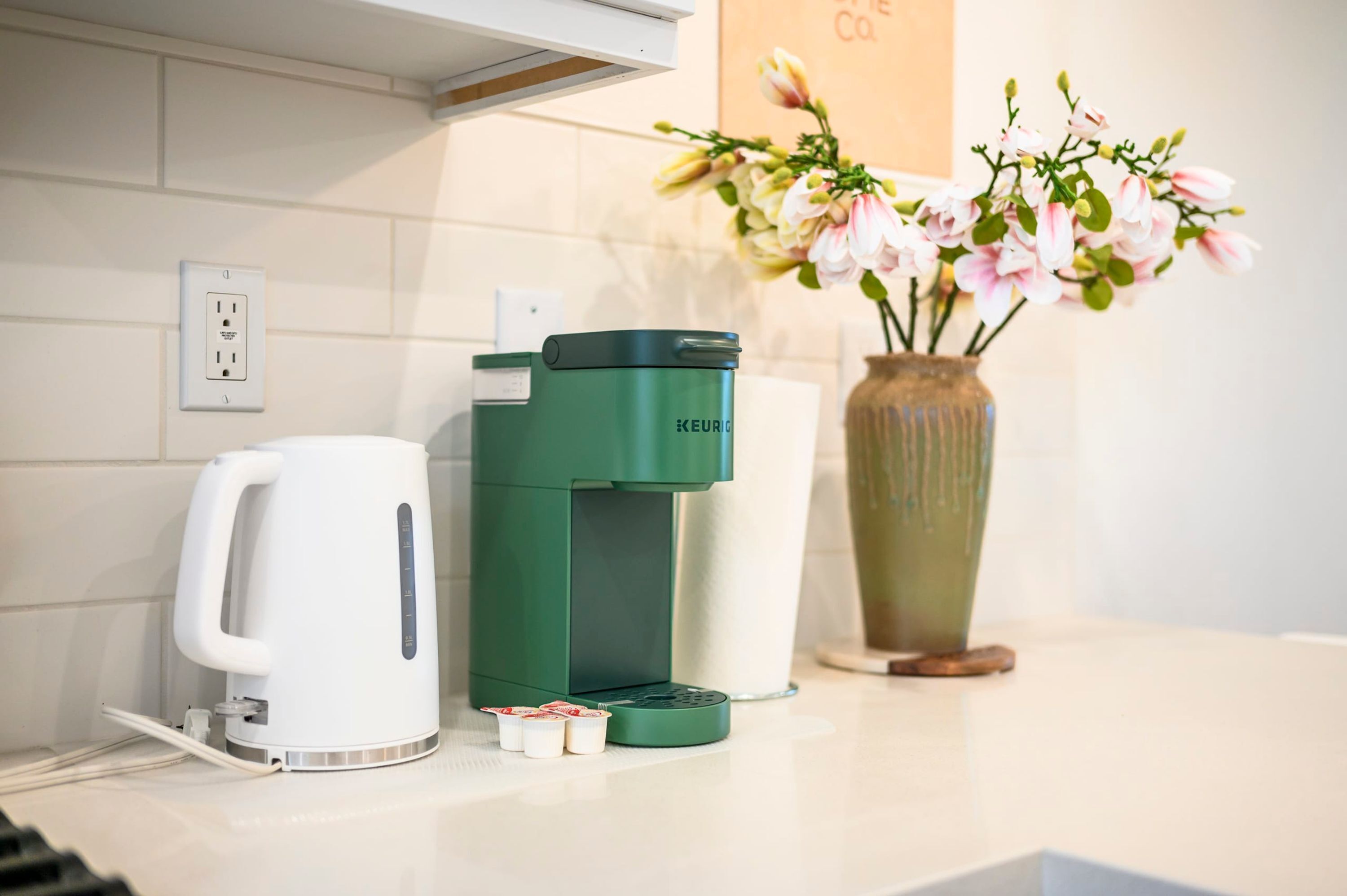 A coffee maker, a white electric kettle, and a vase with flowers on a kitchen counter.