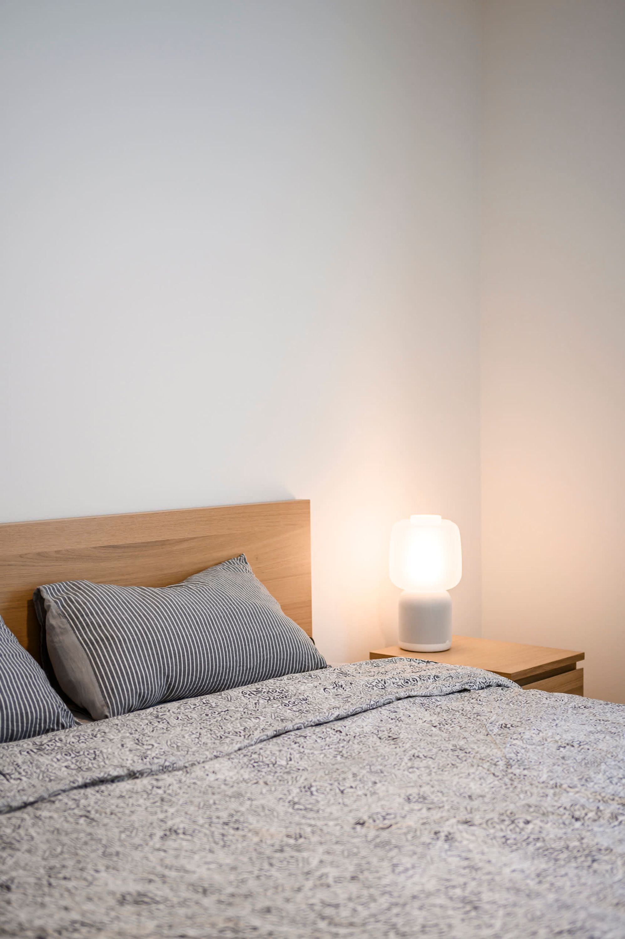 Een net opgemaakt bed met een bedekt en bekleed met kussens, een nachtkastje met een lamp en een houten hoofdbord.