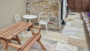 A wooden picnic table with benches, two white metal chairs, and a small round table with a white lace tablecloth.
