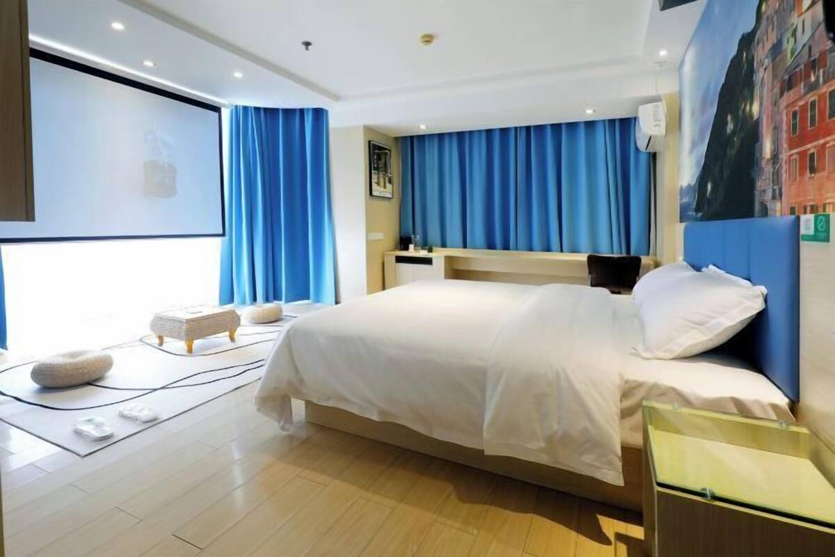 Una habitación de hotel moderna con una cama grande, un televisor de pantalla plana, un sillón de masajes y vistas a un paisaje urbano a través de la ventana.
