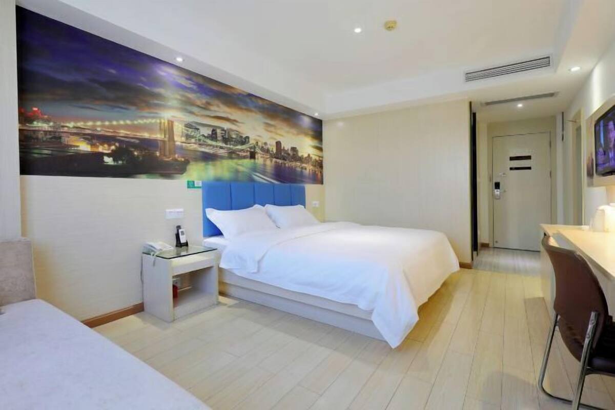 Habitación de hotel con cama, escritorio, silla, mesita de noche y un cuadro grande con una vista urbana.
