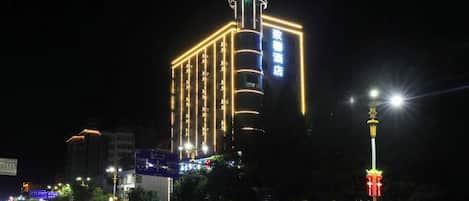 Un hotel de varios pisos con señalización iluminada, una estructura vertical destacada y vista a la calle con una valla y farolas.