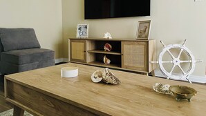 A living room with a flat-screen TV mounted on the wall, a wooden coffee table with decorative shells, a grey sofa, and a white ship's wheel decoration. - Soothing Beach Apartment in Redondo Beach…20 steps to Ocean! (Redondo Beach)