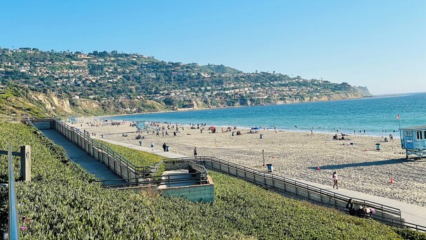 A coastal beach with a walkway, a lifeguard tower, and houses on a hillside in the background. - Soothing Beach Apartment in Redondo Beach…20 steps to Ocean! (Redondo Beach)