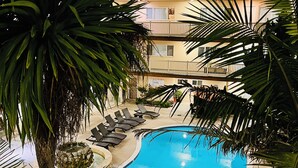 A swimming pool surrounded by lounge chairs and an umbrella, with palm trees and a building in the background. - Soothing Beach Apartment in Redondo Beach…20 steps to Ocean! (Redondo Beach)