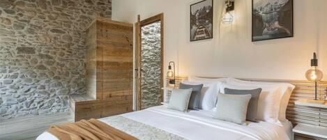 A bedroom with a stone wall, wooden ceiling, a bed with white bedding, two wall-mounted lights, and two framed pictures.