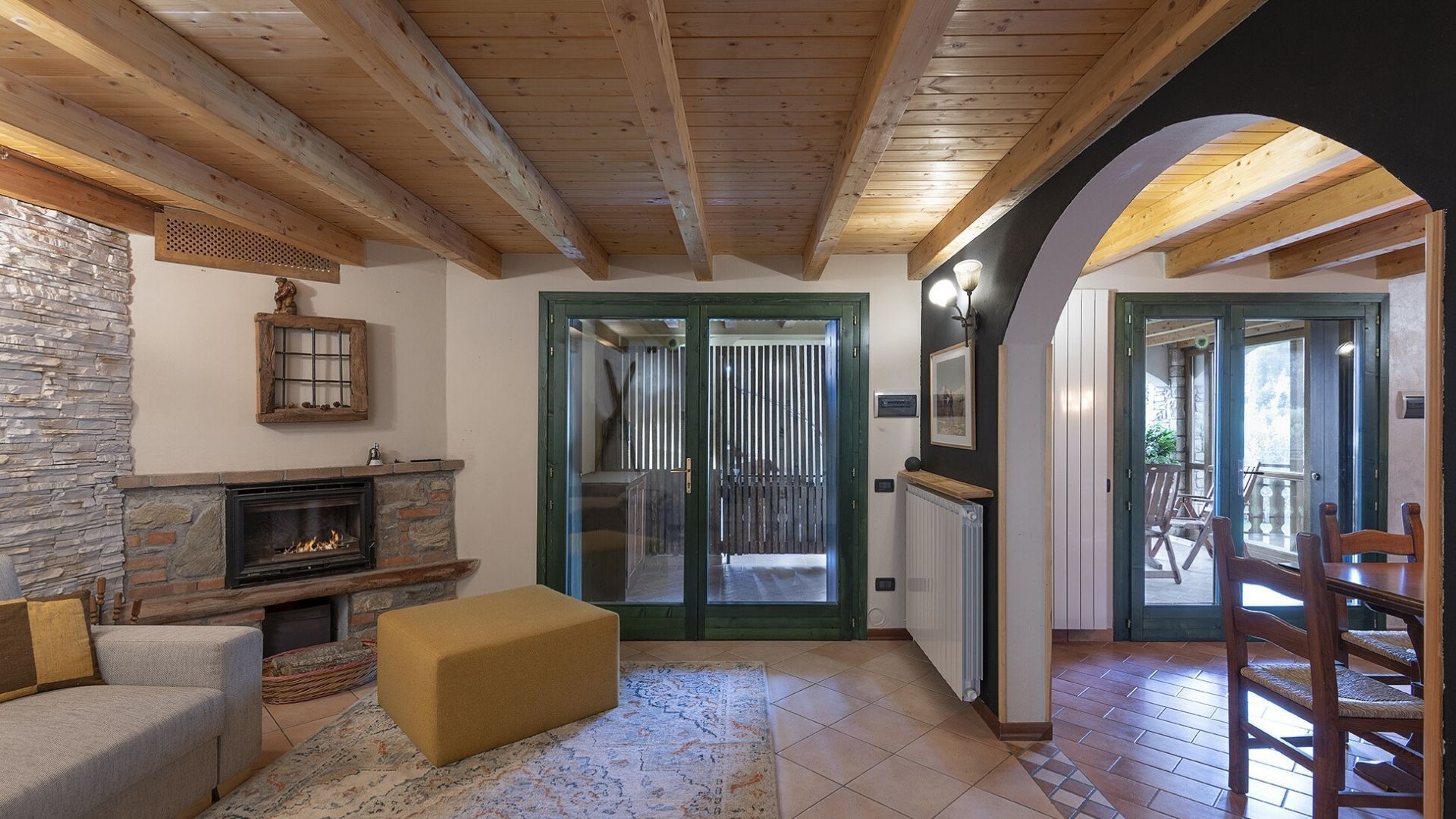 A cozy living room with a fireplace, a stone wall, and a wooden ceiling. There is a door leading to an outdoor area with chairs.