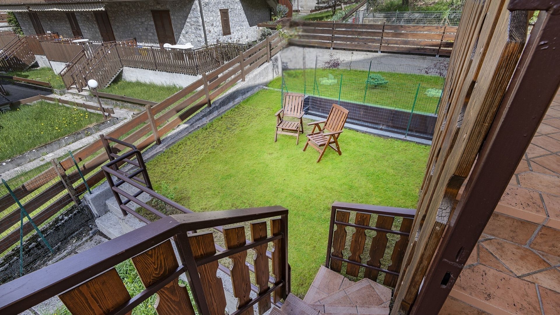 A balcony with a view of a grassy yard, wooden chairs, and a stone building in the background.