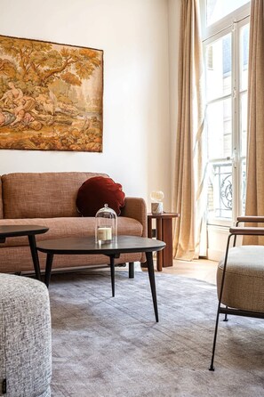 Uma sala de estar com sofá, mesa de centro, mesa lateral e uma cadeira. Há um quadro na parede e uma janela com cortinas.