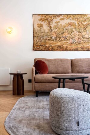 Uma sala de estar moderna com um sofá, um banquinho redondo, uma mesinha e uma grande pintura de paisagem na parede.