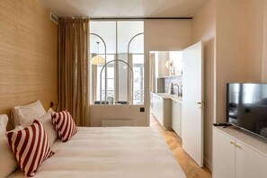 A hotel room with a bed, a TV, a kitchenette, and a window with curtains.