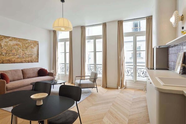A modern living room with a sofa, dining table, and chairs, large windows with curtains, and a kitchen area with a counter and cabinets.