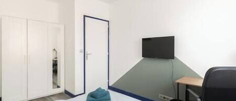 A hotel room with a bed, a desk, a chair, a TV mounted on the wall, and a wardrobe.