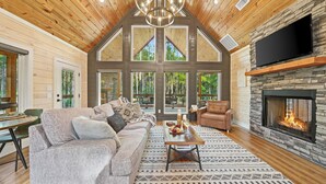 A cozy living room with a stone fireplace, a large TV mounted on the wall, and a high ceiling with wooden beams. - Romantic Cabin Swings | Private Sauna & Hot Tub! (Broken Bow)