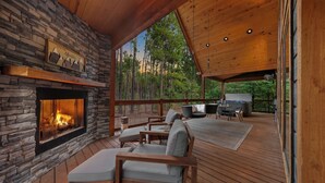 A cozy outdoor deck with a stone fireplace, comfortable seating, and a view of the forest. - Romantic Cabin Swings | Private Sauna & Hot Tub! (Broken Bow)