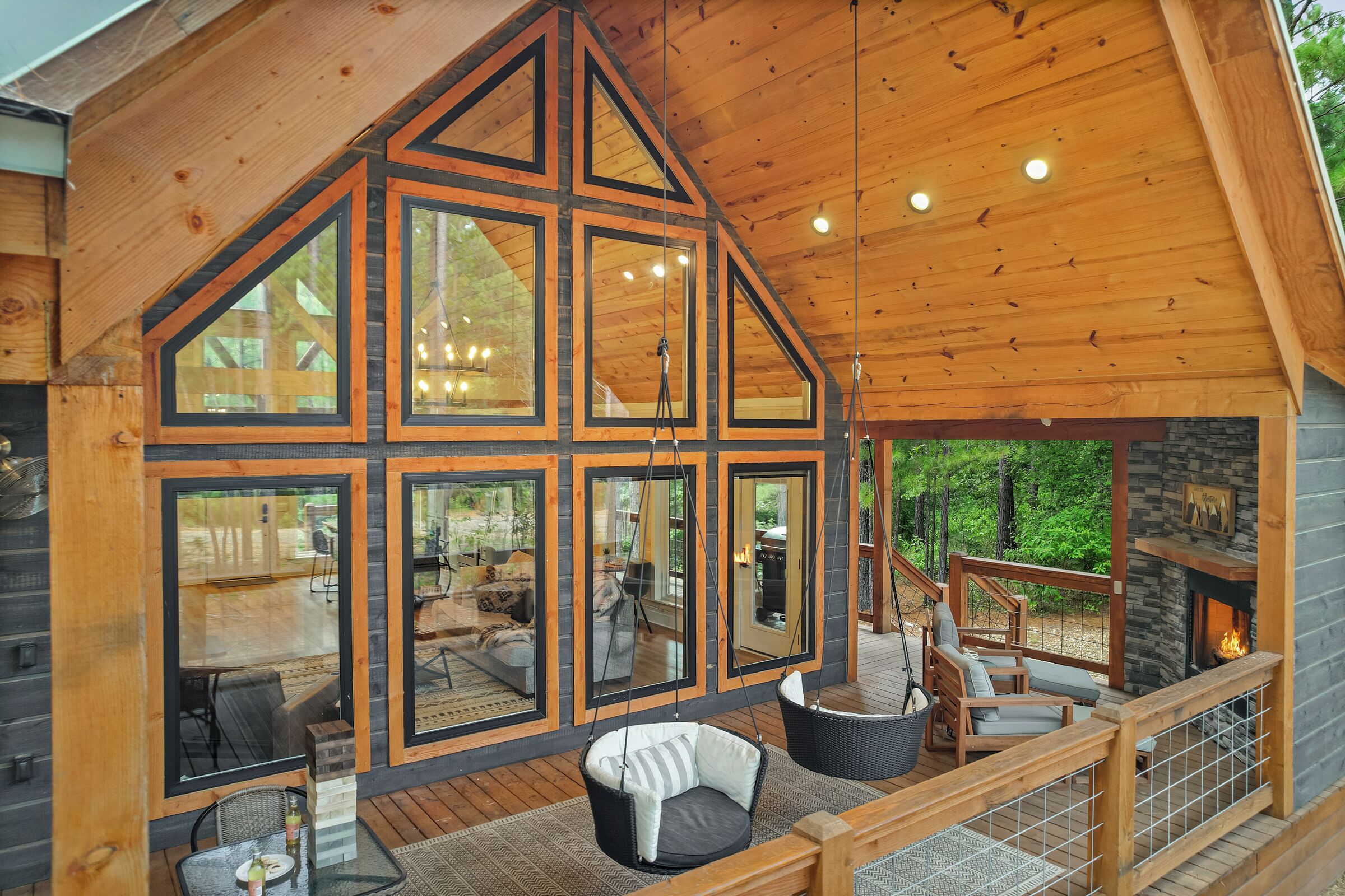 A cabin with large windows, a wooden deck, a hanging chair, and a fireplace.