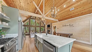 A spacious kitchen with a large island, wooden ceiling beams, and a stone fireplace. - Romantic Cabin Swings | Private Sauna & Hot Tub! (Broken Bow)