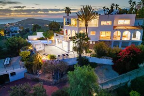 Una casa moderna de dos pisos con balcón, rodeada de abundante vegetación y vista al mar.