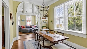 A dining area with a wooden table and chairs, a chandelier, and a view of the outdoors through large windows. - Bamboo & Clover - Historic Galveston Cottage (Galveston)