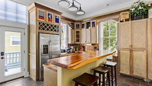 A kitchen with wooden cabinets, a stainless steel refrigerator, a large island with stools, and a sign that says "SHARE THE PIE". - Bamboo & Clover - Historic Galveston Cottage (Galveston)