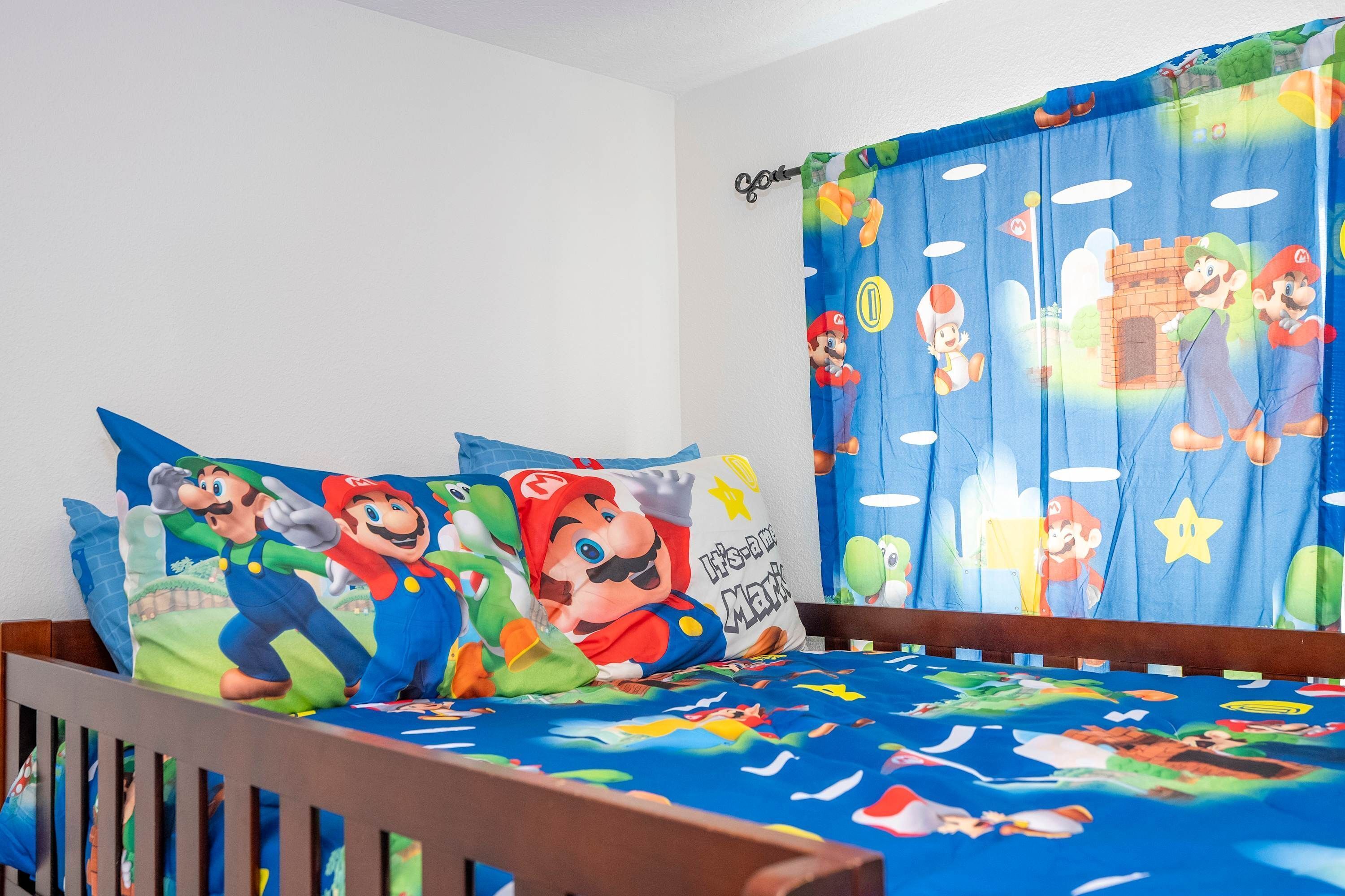 A bedroom with a bed covered in Mario-themed bedding, a pillow with Mario and Luigi, and a window with a Mario curtain.