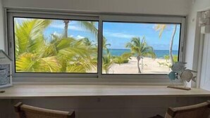 Ein Zimmer mit Schreibtisch und zwei Korbstühlen, ein Fenster mit Blick auf Palmen und Strand.