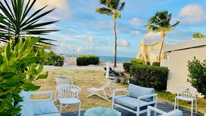 A beachside patio with white lounge chairs and a small table, palm trees, and a view of the ocean. - Côté Mer - NBBC - 2 bedrooms beachfront (Marigot)