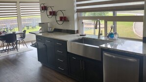 A modern kitchen with dark cabinetry, a stainless steel sink, and a dishwasher. There are two windows with blinds, and a dining area with a table and chairs is visible in the background. - 2-bedroom, pet friendly home with great amenities. It will be your happy place (Holly Lake Ranch)