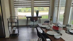 A dining area with a long wooden table, black chairs, and a white rug. There are windows with blinds, a potted plant, and a view of a green landscape. - 2-bedroom, pet friendly home with great amenities. It will be your happy place (Holly Lake Ranch)