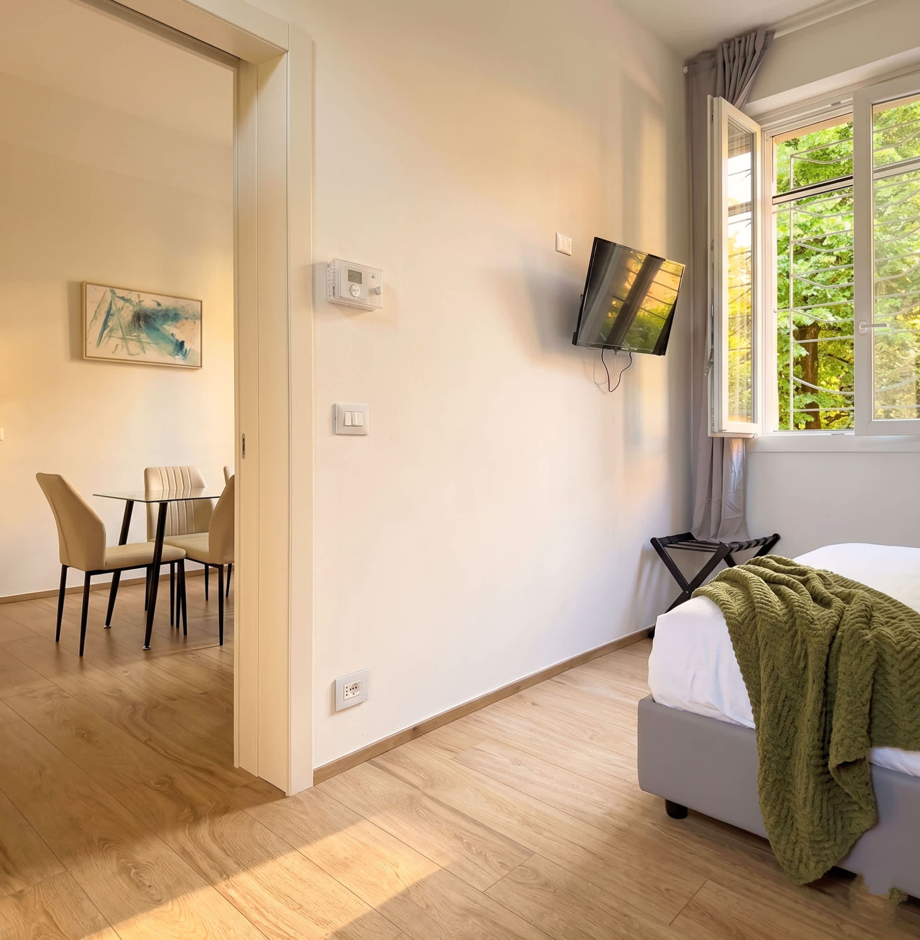 A hotel room with a bed, a TV mounted on the wall, a window with a view of trees, a painting on the wall, and a dining area with a table and chairs.
