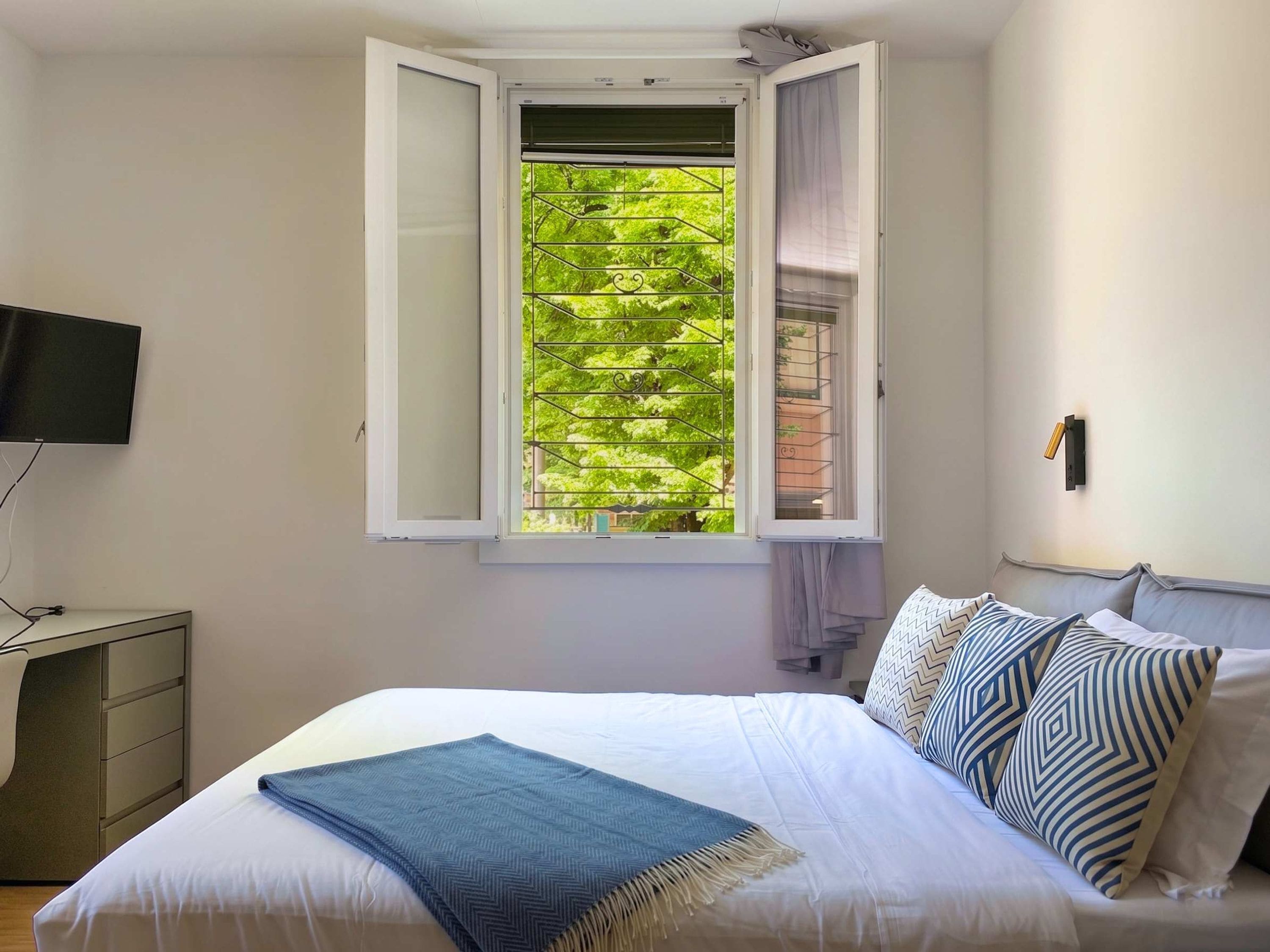 A bedroom with a bed, a desk, a TV, and a window with an open shutter.