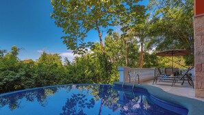 Una piscina cristallina blu circondata da vegetazione lussureggiante, con una zona patio arredata con mobili da esterno e un muro in pietra.