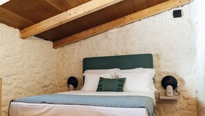 Une chambre avec un mur en pierre, un plafond en bois, un lit avec une literie blanche, une tête de lit verte et deux lampes de chevet.