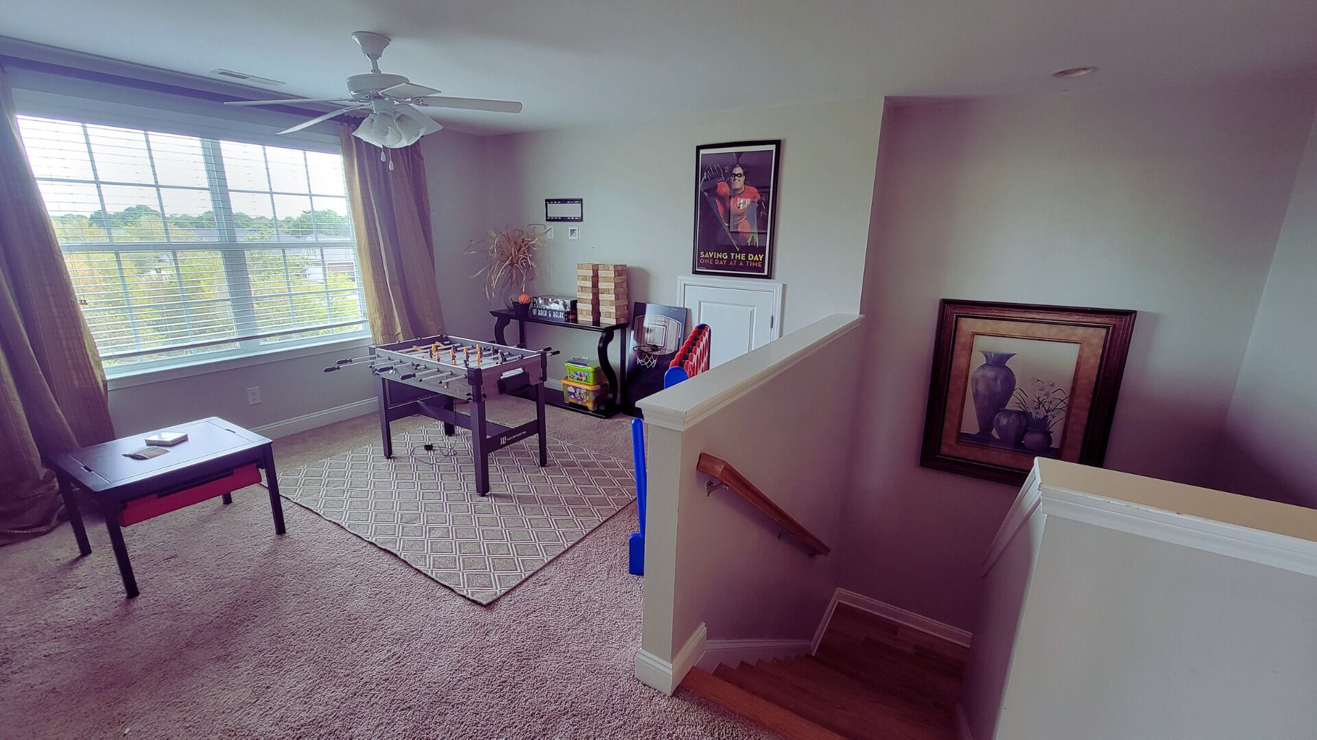 A living room with a foosball table, a ceiling fan, a window with curtains, and a framed picture on the wall.