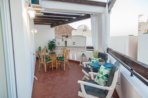 Una terrazza esterna con un tavolo rotondo in vetro e sedie in vimini, un piccolo tavolo rotondo con una tovaglia blu e una ringhiera bianca.