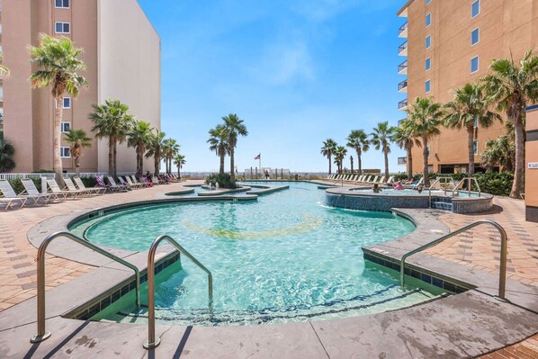 A swimming pool with multiple lanes, surrounded by palm trees and lounge chairs, with a hotel building in the background. - Crystal Tower 206- Amenities Galore Lazy River (Gulf Shores)