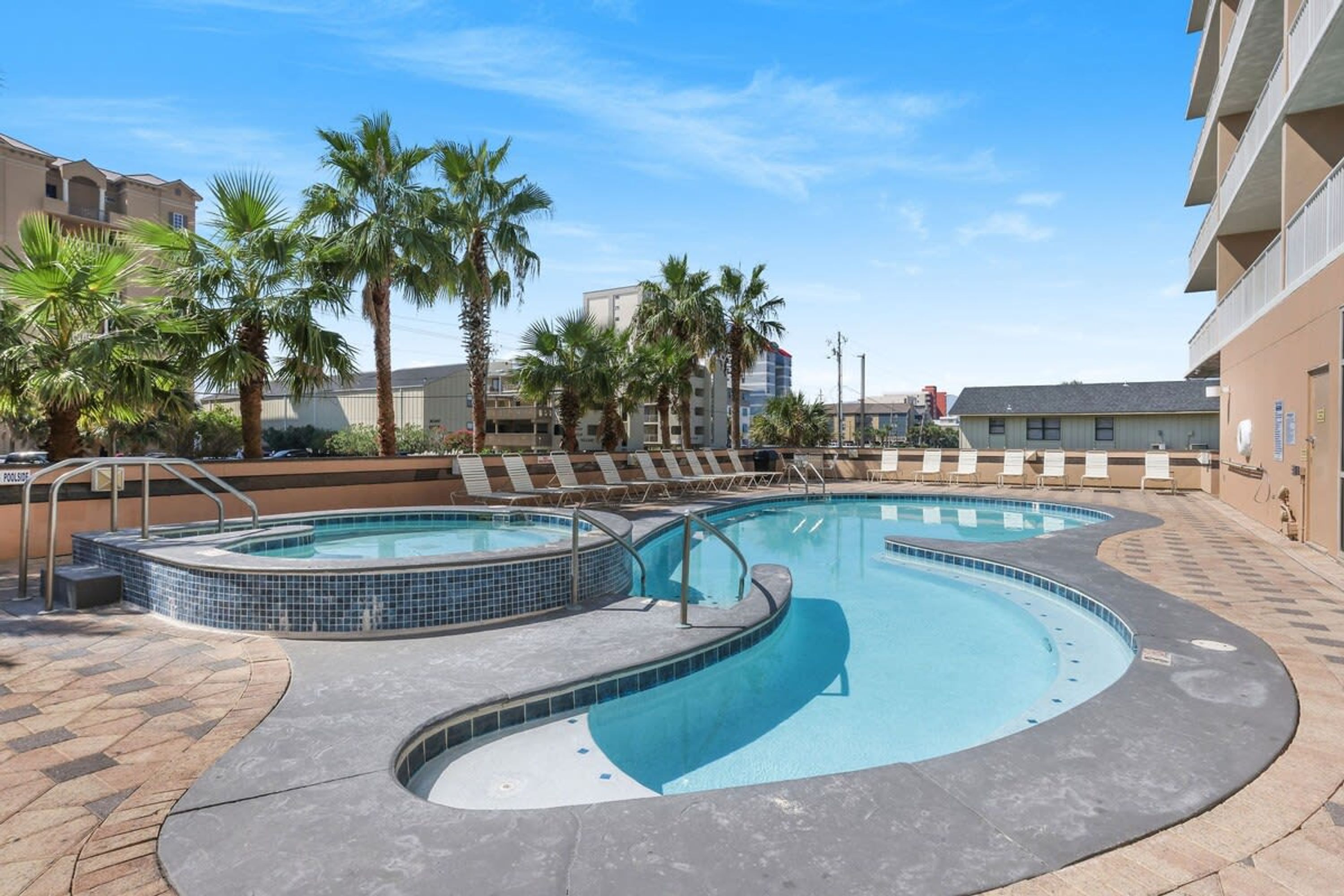 A swimming pool area with two pools, palm trees, lounge chairs, and a tiled deck.