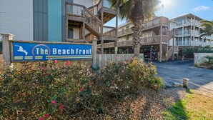 Un hotel frente a la playa con un letrero que dice "The Beach Front", con una escalera de madera que conduce a un balcón, rodeado de vegetación exuberante y una palmera.