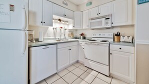 Una cocina compacta con armarios blancos, microondas, estufa y lavavajillas. Hay letreros en los armarios y una pequeña ventana sobre el fregadero.