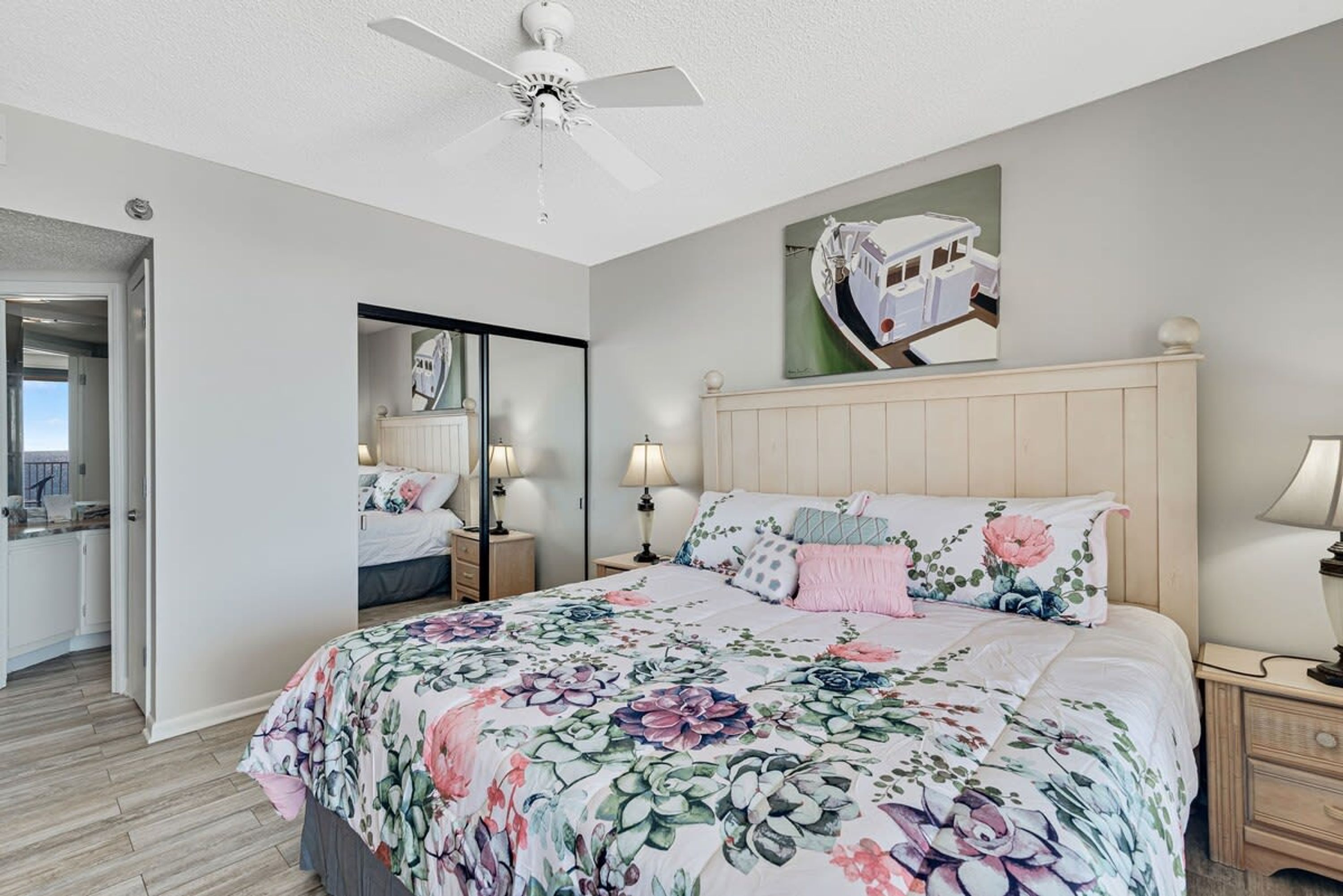 A bedroom with a floral bedspread, a ceiling fan, a nightstand with lamps, and a mirror on the wall.