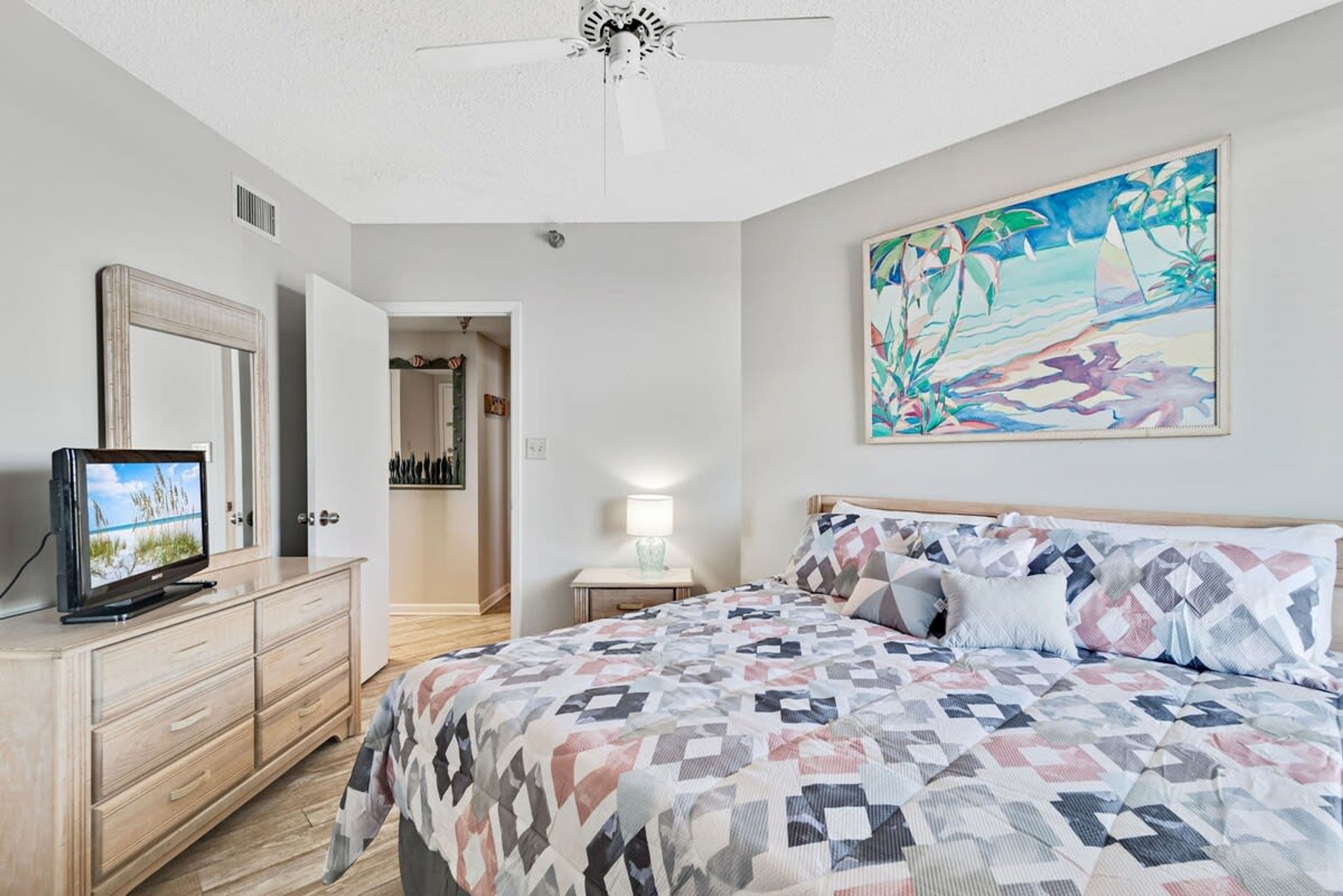 A bedroom with a bed, a TV on a dresser, a painting on the wall, and a door leading to another room.
