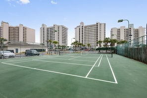 Ein Außen-Tennisplatz mit Netz, umgeben von einer Umzäunung, neben einem Parkplatz.