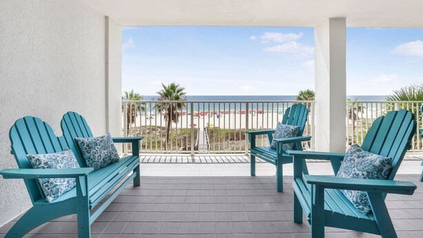 A balcony with four turquoise Adirondack chairs, each with a patterned cushion, overlooking a beach with palm trees and a clear sky. - Windward Pointe 303 Ideally Located on 3rd Floor (Orange Beach)