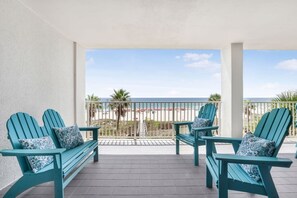 A balcony with four turquoise Adirondack chairs, each with a patterned cushion, overlooking a beach with palm trees and a clear sky. - Windward Pointe 303 Ideally Located on 3rd Floor (Orange Beach)