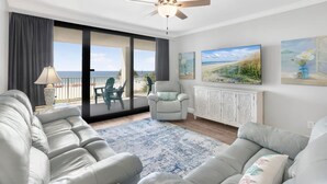 A spacious living room with a large window, a ceiling fan, a gray sofa, a white cabinet, and a blue and white rug. - Windward Pointe 303 Ideally Located on 3rd Floor (Orange Beach)