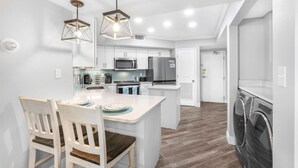 A modern kitchen with white cabinetry, a central island, and a dining area with two chairs. - Windward Pointe 303 Ideally Located on 3rd Floor (Orange Beach)