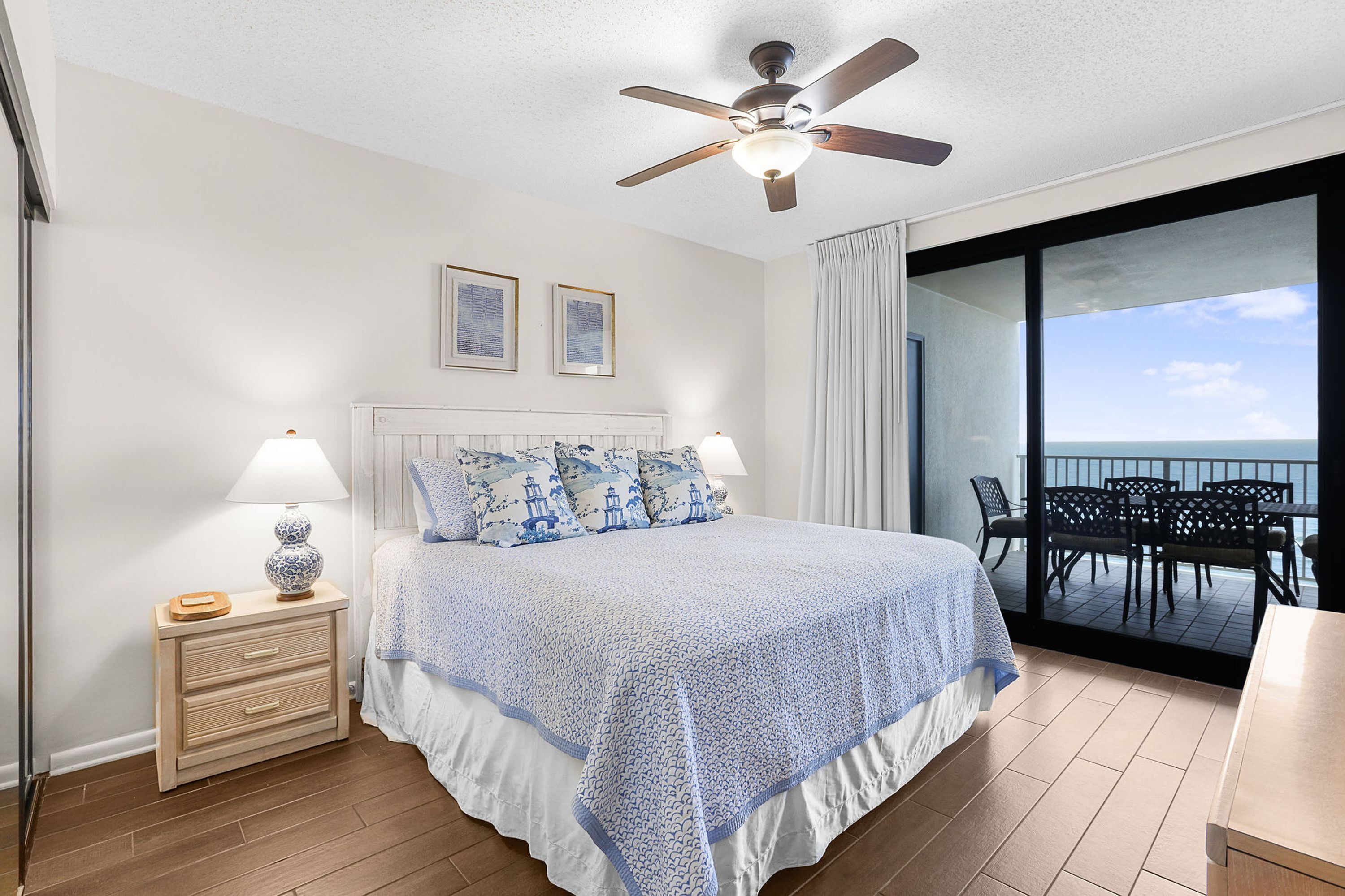A bedroom with a bed, bedside lamps, a ceiling fan, and a view of the ocean through a balcony.